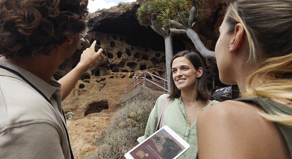 Dos personas observando y señalando las cuevas del Cenobio de Valerón, con una tableta en la mano y vegetación en la parte superior de la formación rocosa. Dos personas observando y señalando las cuevas del Cenobio de Valerón, con una tableta en la mano y vegetación en la parte superior de la formación rocosa.